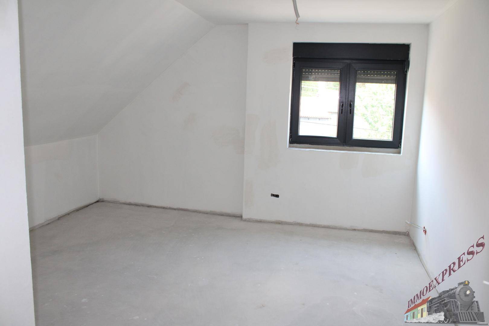 Unfertiger Raum im Dachgeschoss mit schrägen Wänden und Fenster mit Rollladen.