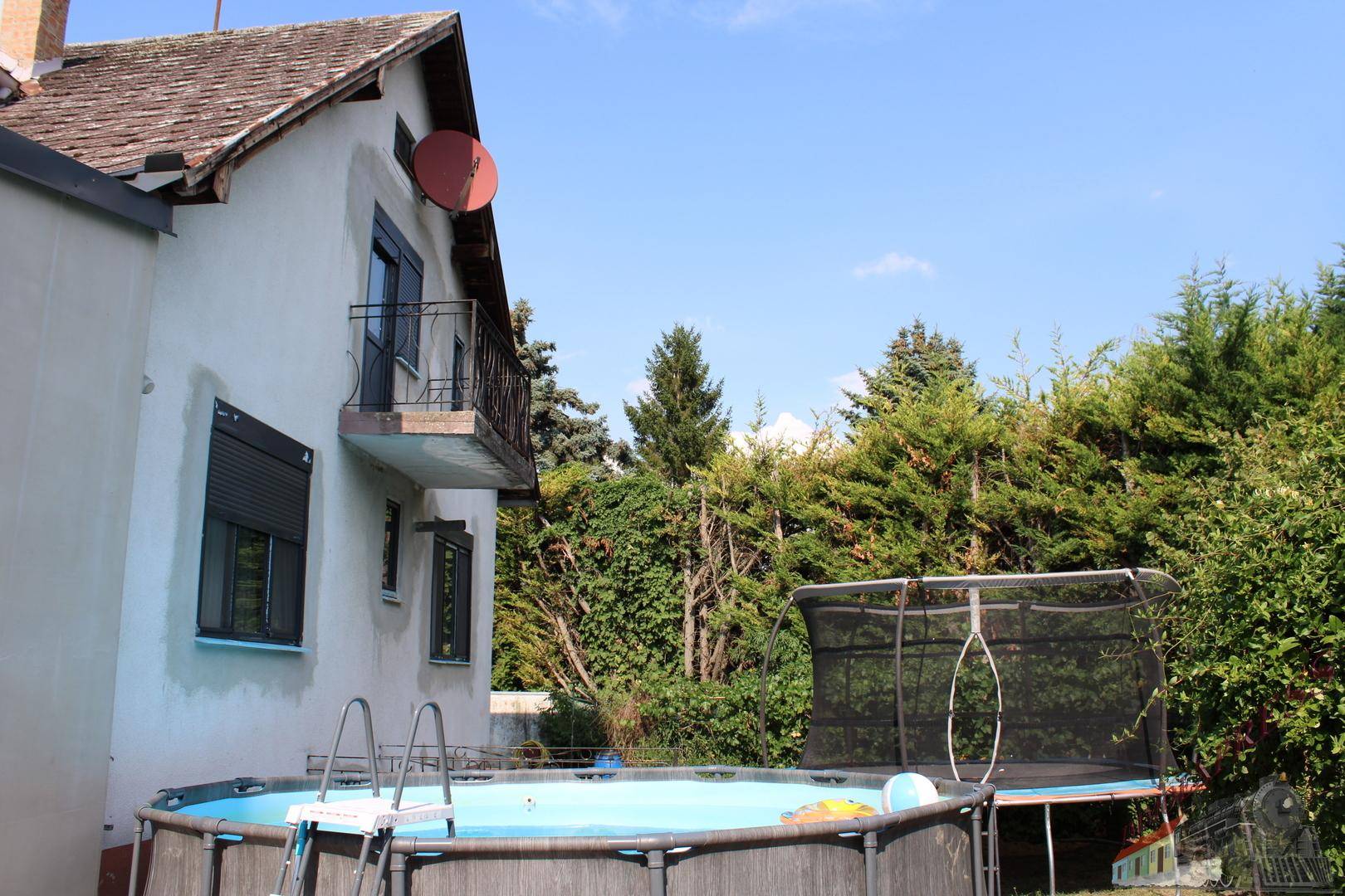 Hausfassade mit Aufstellpool und Trampolin im grünen Gartenbereich.