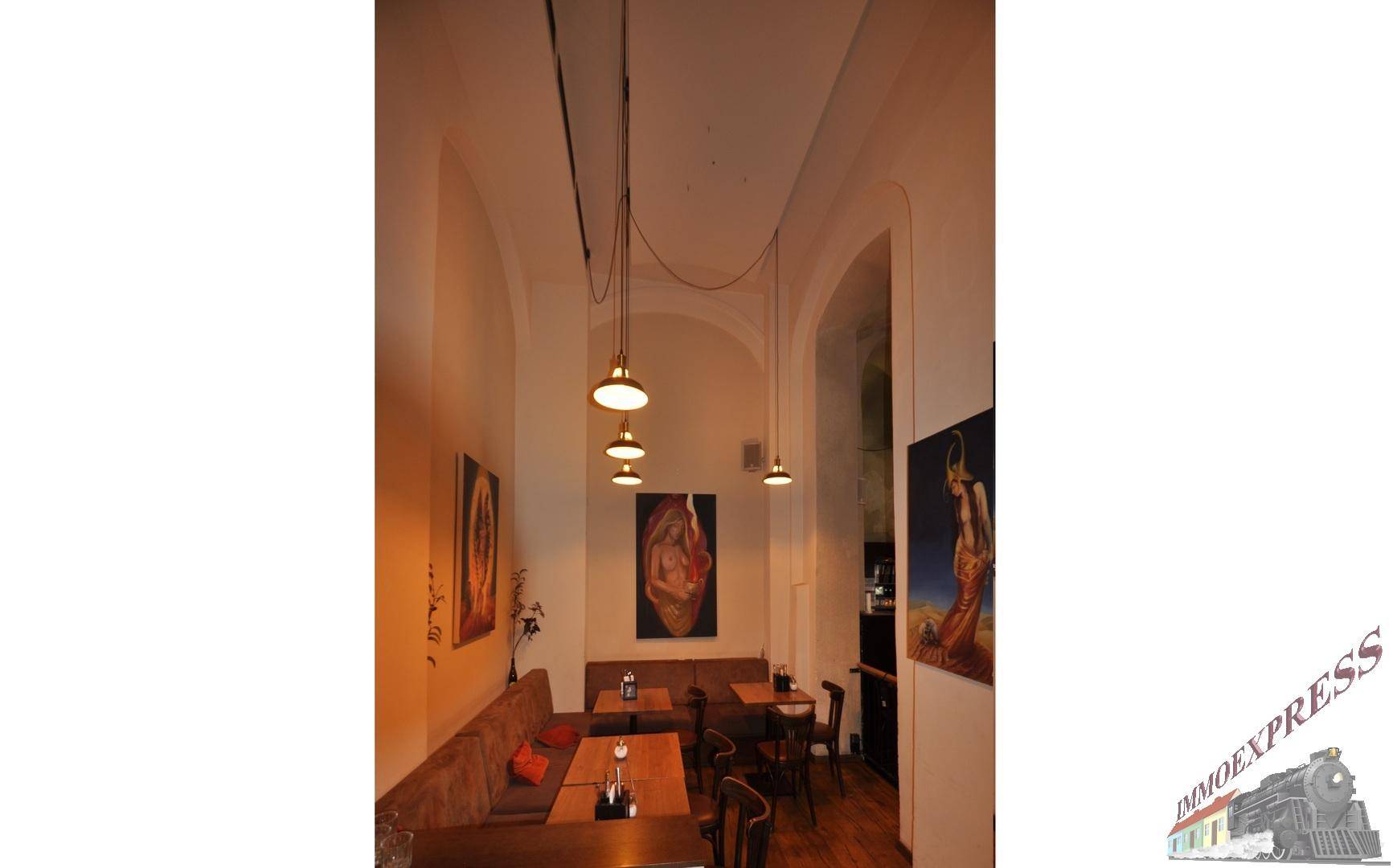 Schmaler Restaurantbereich mit bequemen Sitzbänken, kleinen Tischen und dekorativen Gemälden an den hohen Wänden.
