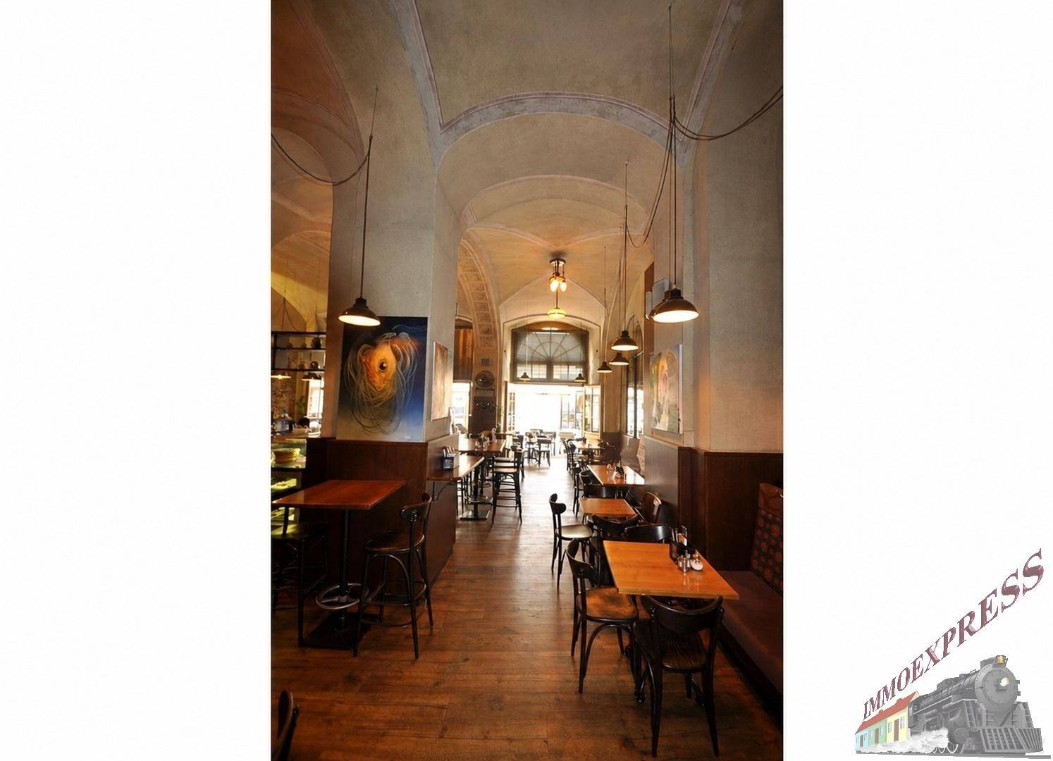 Langer Restaurantflur mit Holzböden, Reihen von Tischen und Stühlen, der zu einem hellen Eingangsbereich führt.