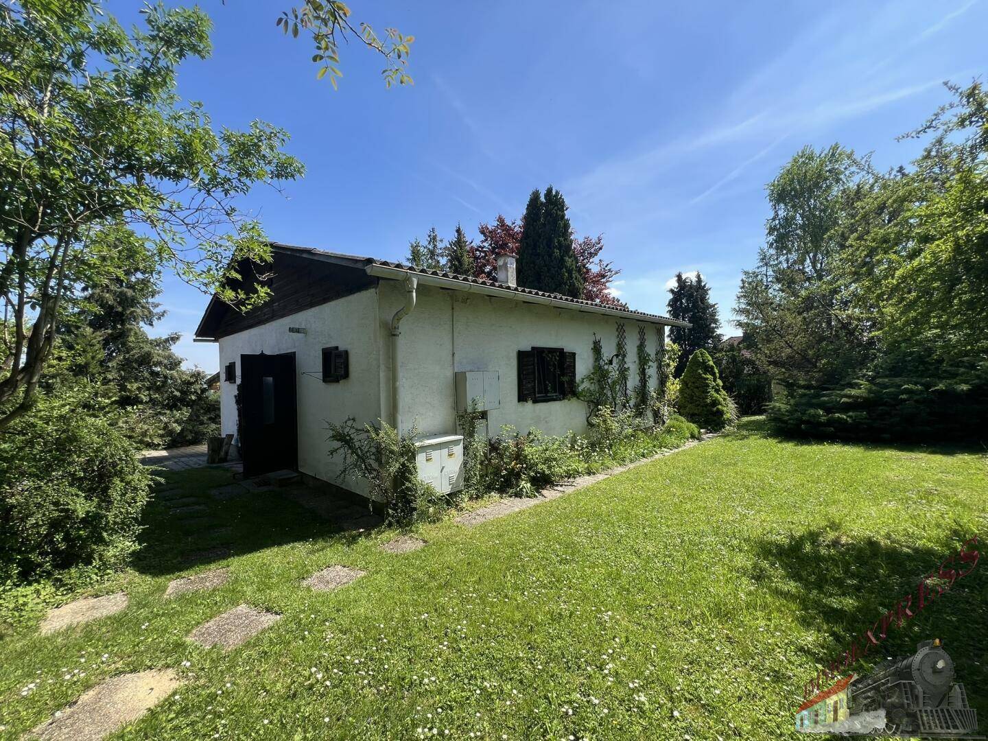 Seitenansicht des Hauses mit gepflegtem Rasen und umgebenden Bäumen unter blauem Himmel.