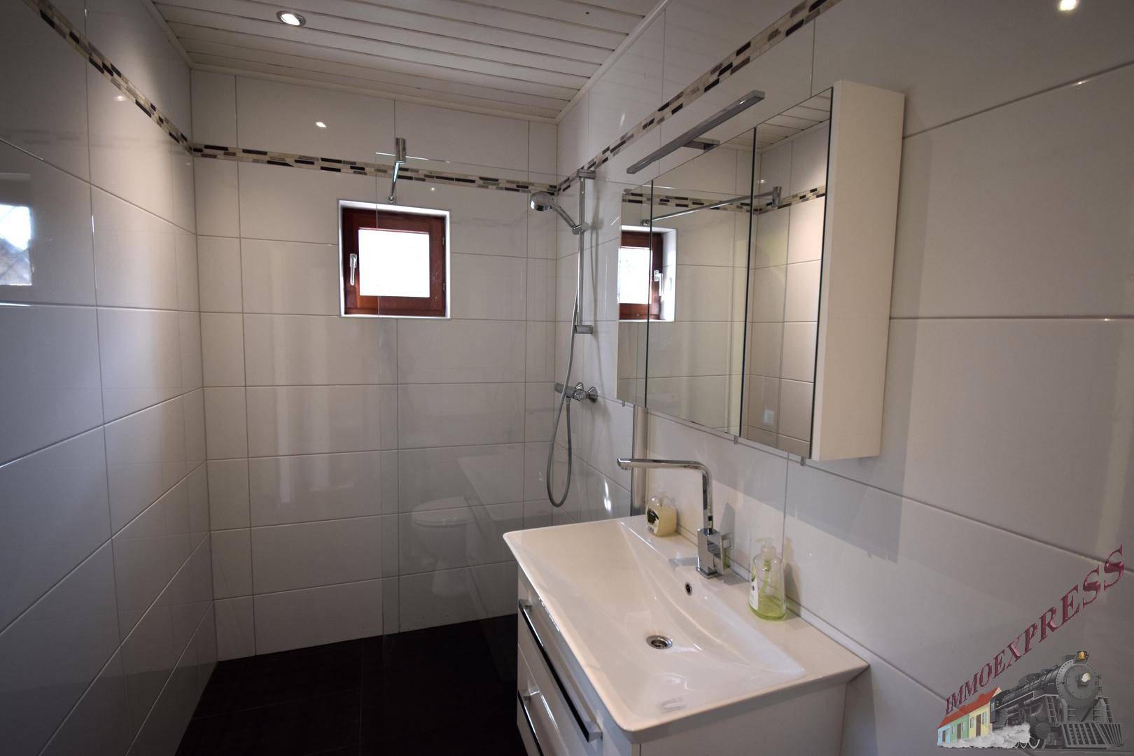 Helles Badezimmer mit weißen Fliesen, großer Dusche und modernem Waschtisch, inklusive Fenster.