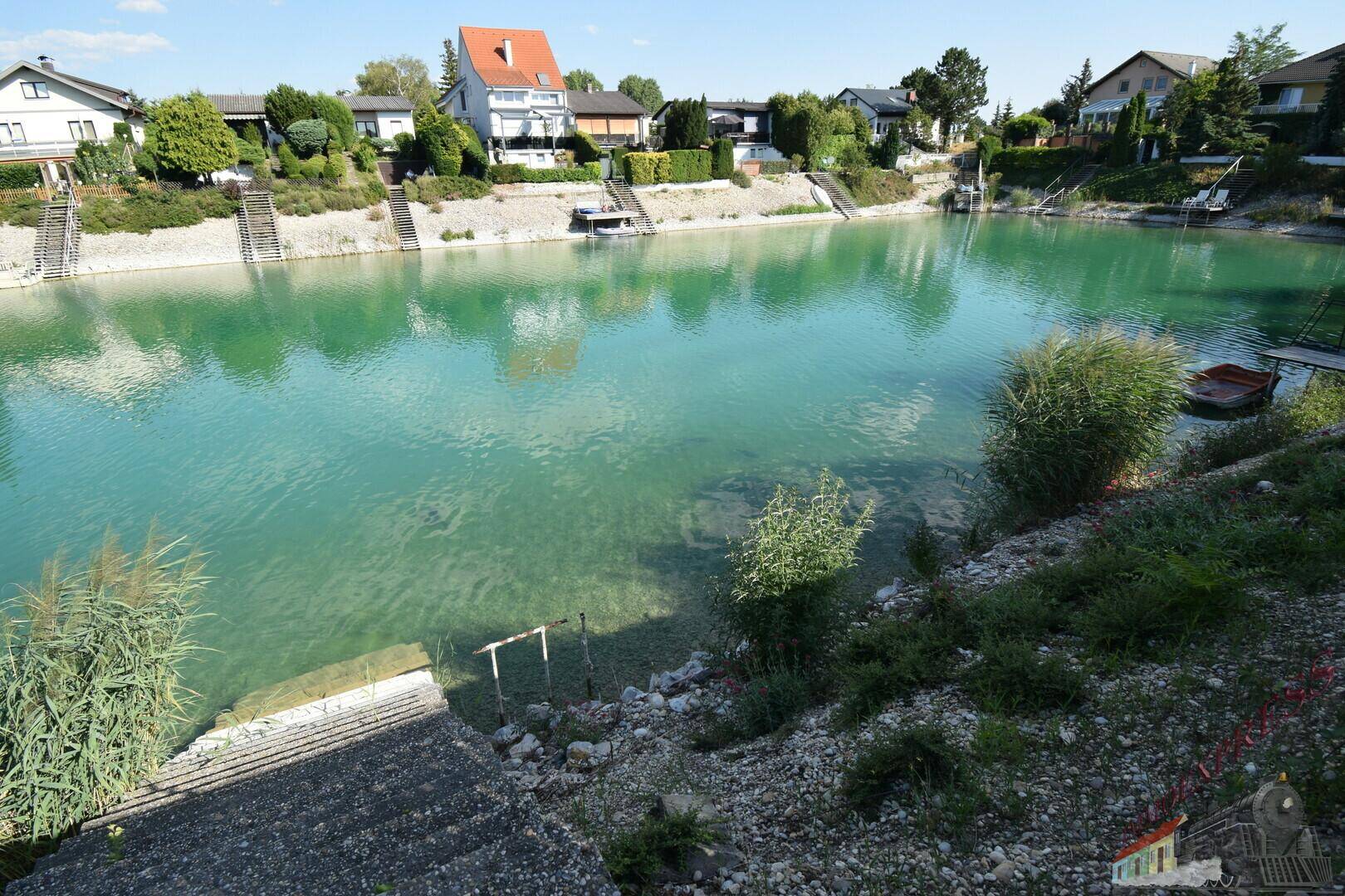 Panoramablick auf den klaren See mit flachem Ufer und Häusern in der Ferne unter strahlendem Sonnenschein.