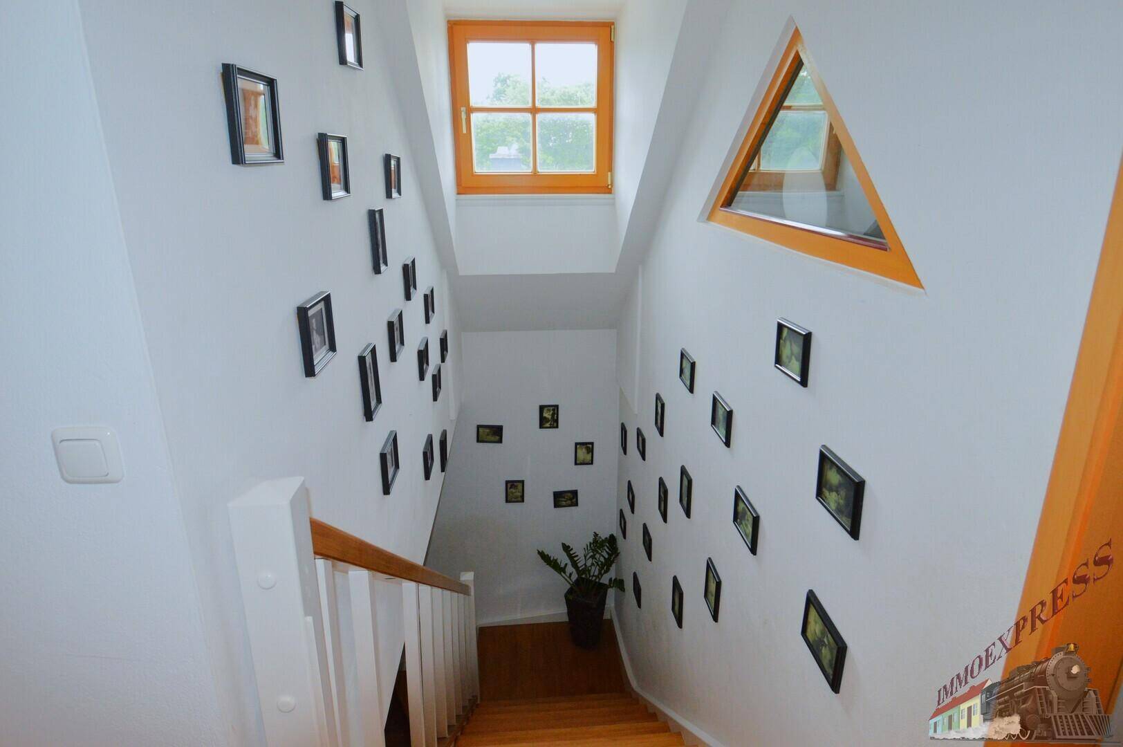 Helles Treppenhaus mit Holztreppe, vielen Bildern und Dachfenster, das viel Licht spendet.