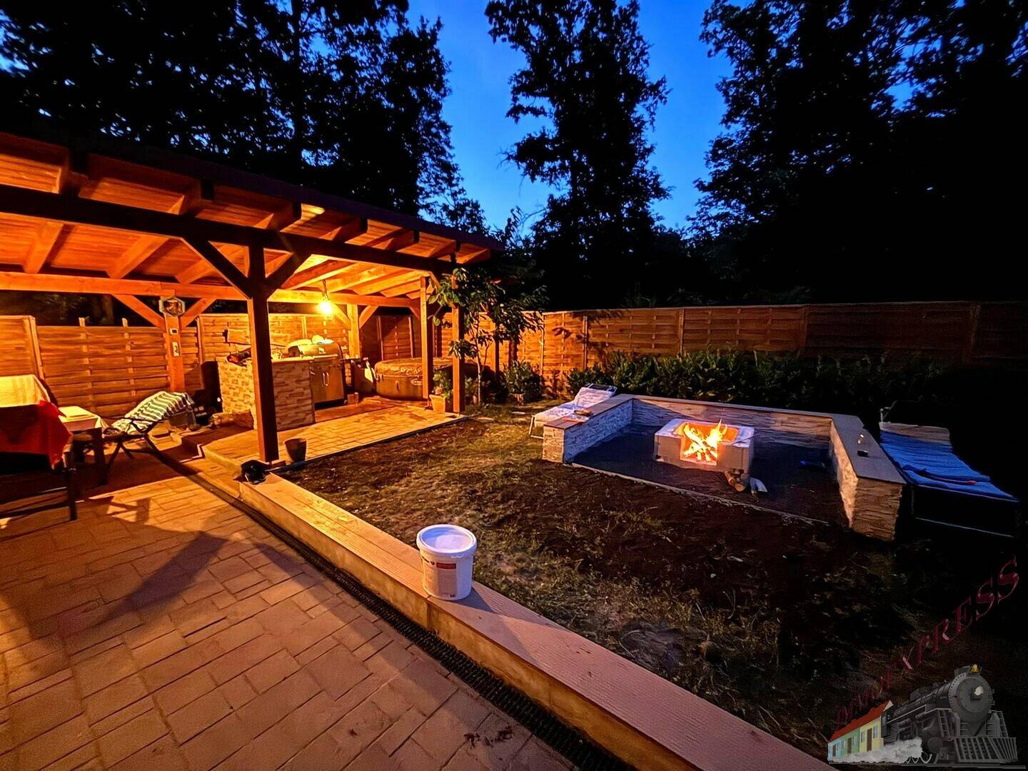 Beleuchteter Gartenbereich bei Nacht mit überdachter Terrasse und brennender Feuerstelle.