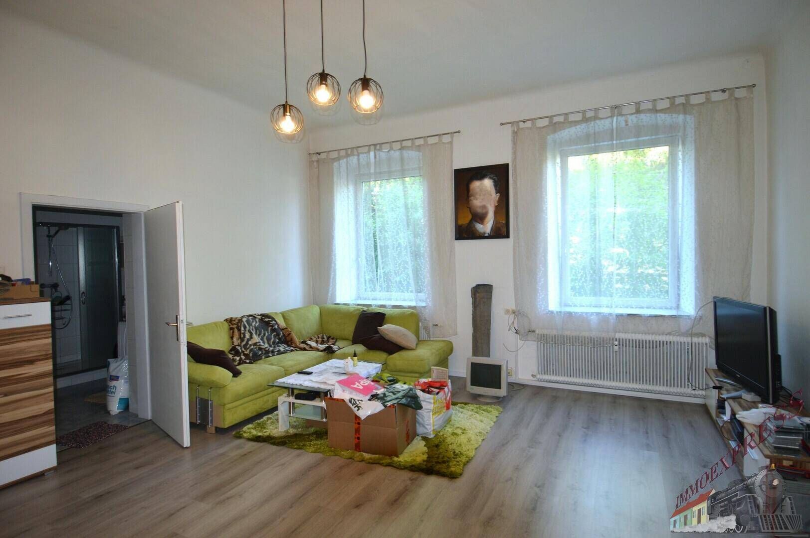 Geräumiges Wohnzimmer mit grünem Ecksofa, zwei Fenstern und Laminatboden.