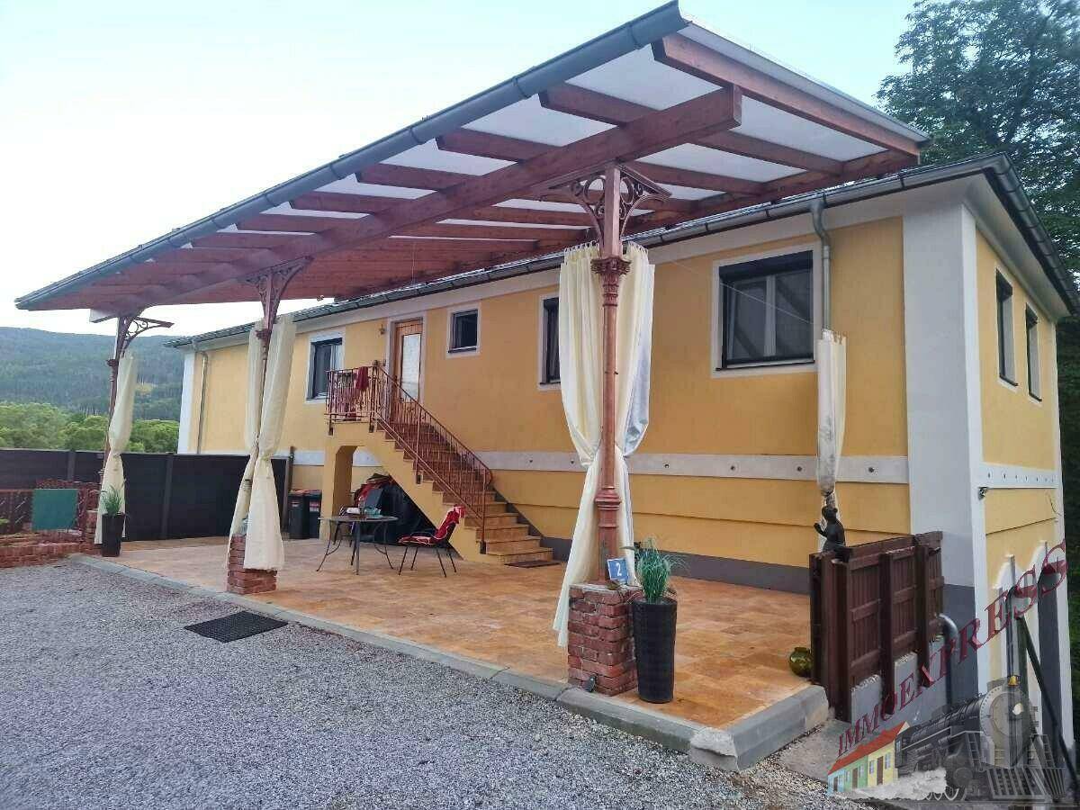 Gelbes Gebäude mit großer Holzpergola über einer gepflasterten Fläche und einer Außentreppe.