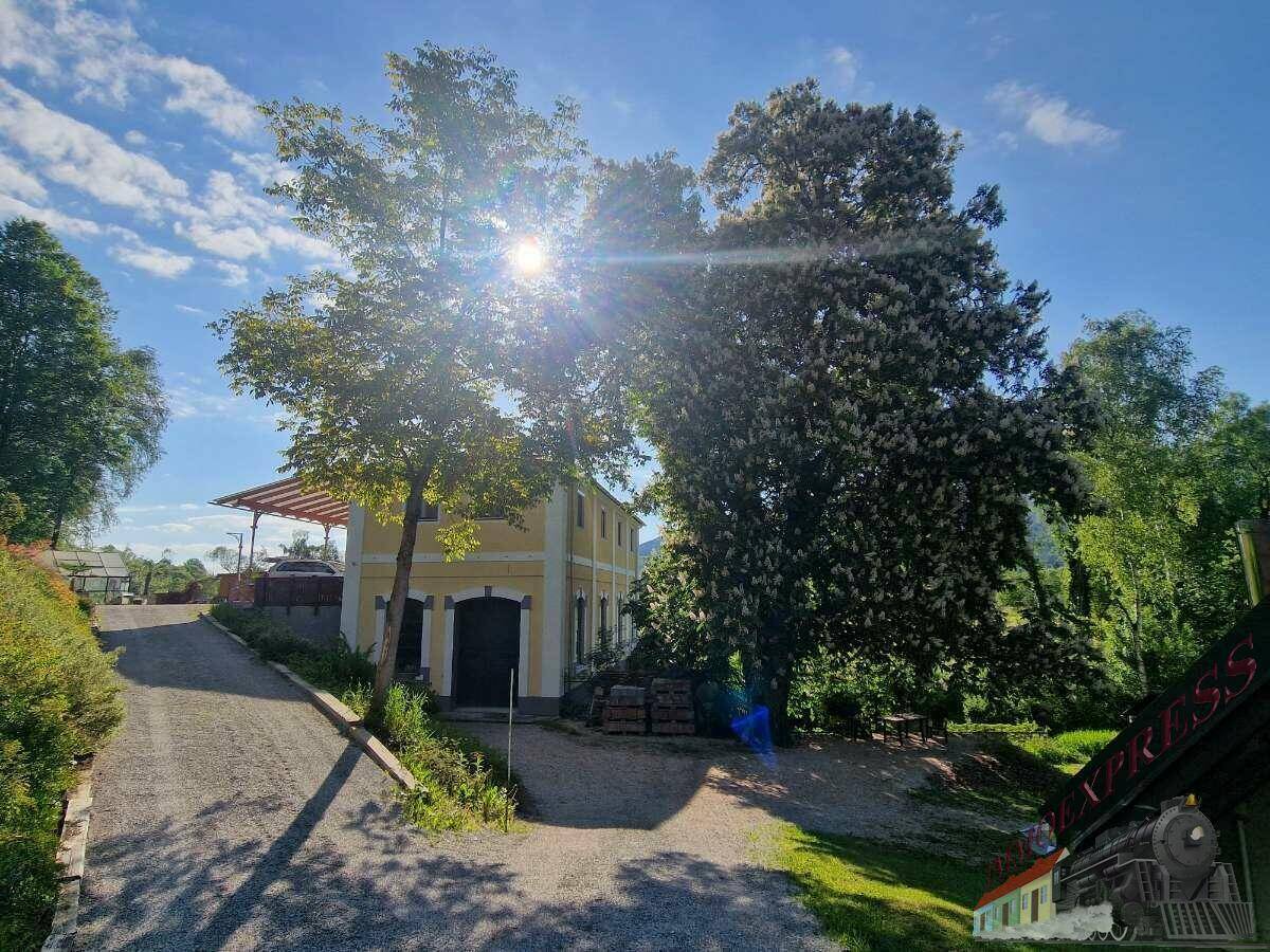 Gelbes Gebäude mit überdachtem Außenbereich, Schotterweg und Bäumen unter strahlendem Sonnenschein.