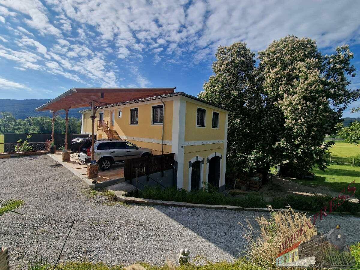 Gelbes Gebäude mit überdachtem Parkplatz und großen Bäumen, umgeben von grüner Landschaft unter blauem Himmel.