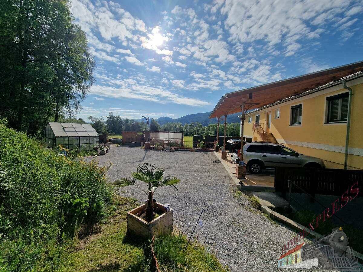 Weitläufiger Kiesparkplatz vor dem gelben Gebäude mit einem Gewächshaus und Bergblick im Hintergrund.