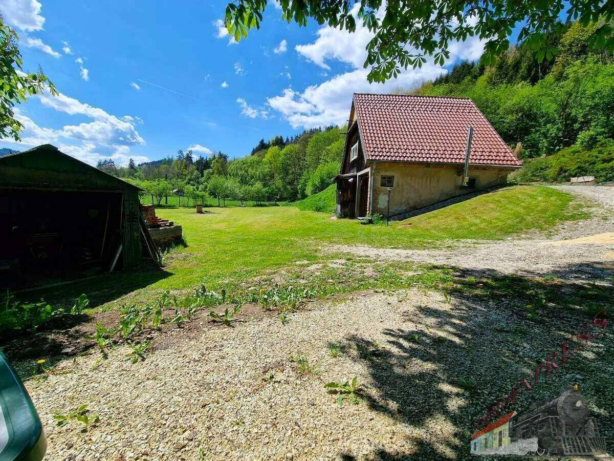Kleines Haus mit Ziegeldach und einem Schuppen auf einer grünen Wiese, umgeben von Bäumen und Natur.