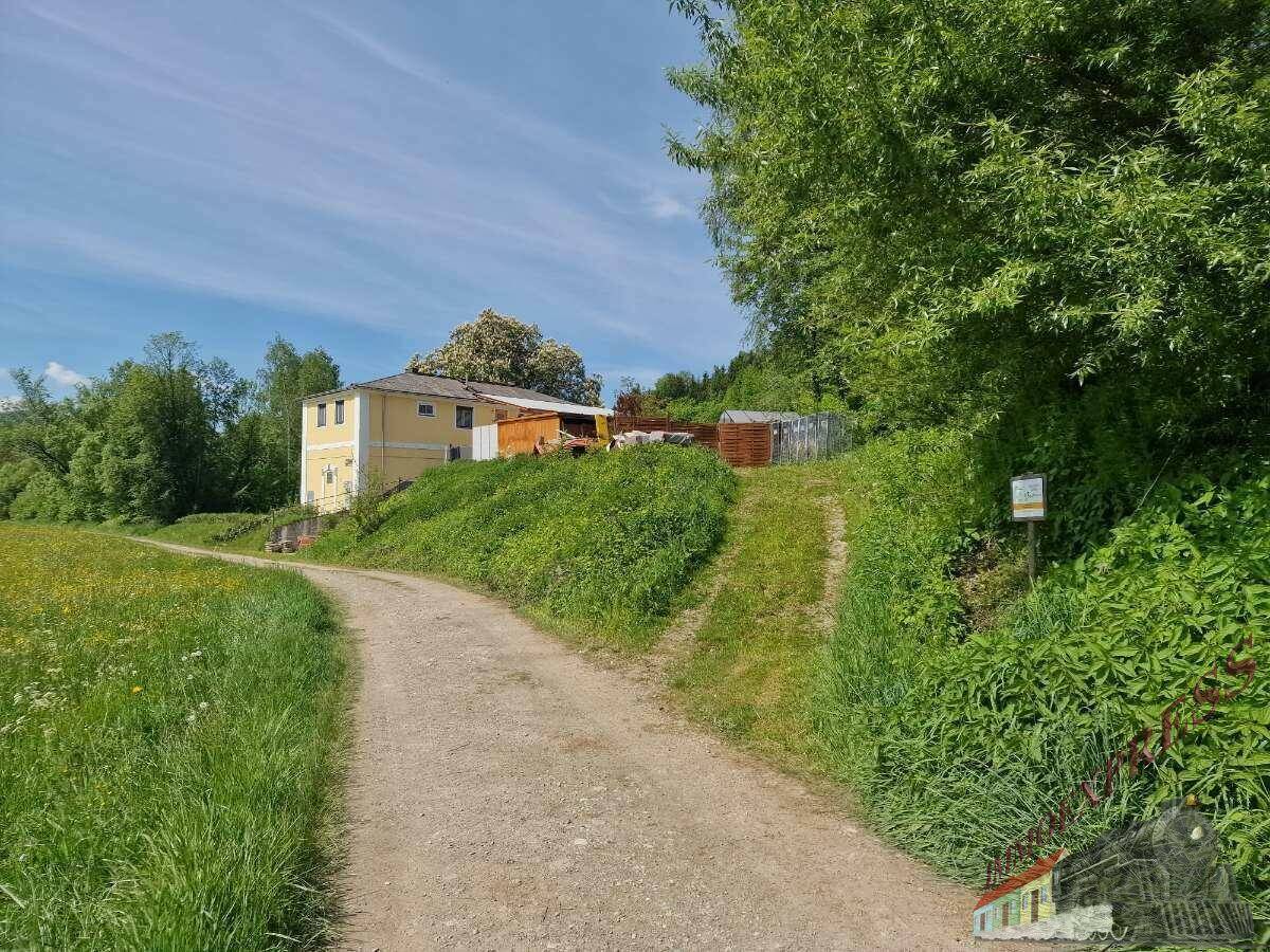 Feldweg führt zu einem gelben Gebäude auf einem Hügel, umgeben von grünen Wiesen und Bäumen.