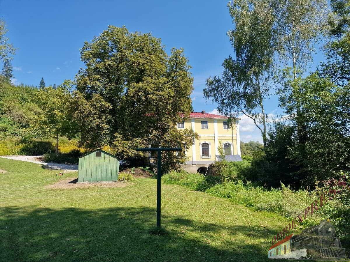 Weitläufige Rasenfläche mit grünem Geräteschuppen und dem gelben Gebäude im Hintergrund, umgeben von Bäumen.