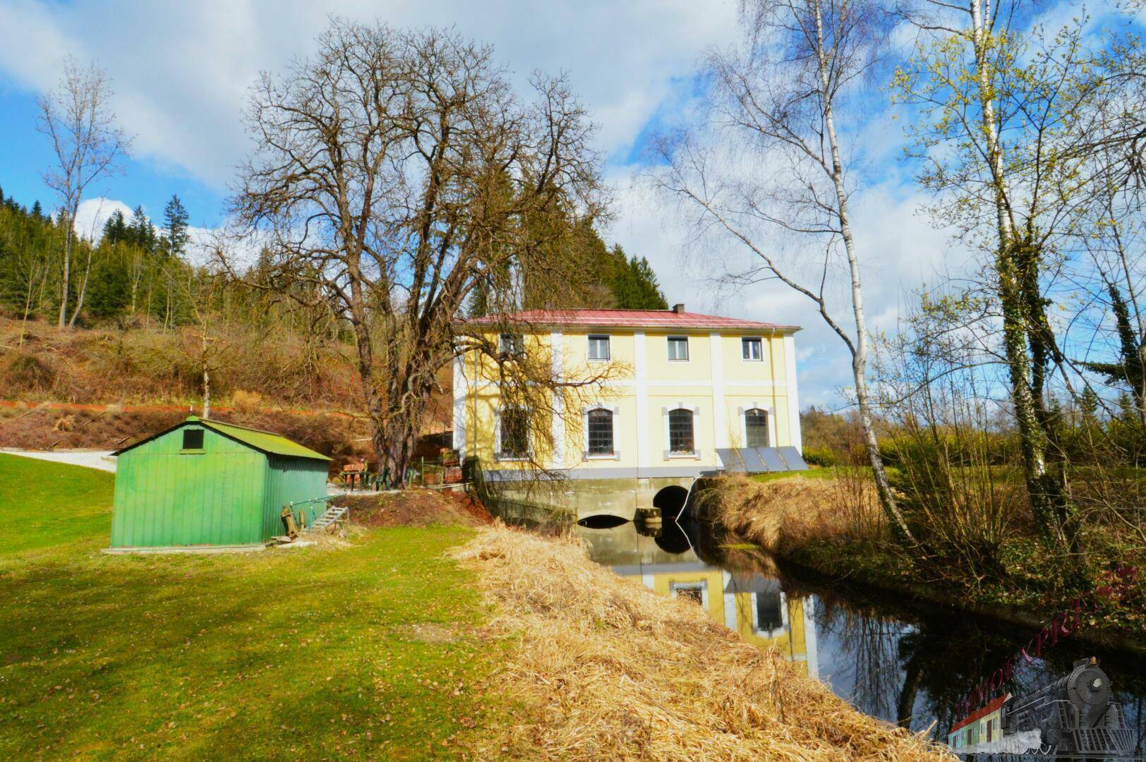 Historisches gelbes Gebäude am Flussufer mit einem grünen Schuppen und üppiger Vegetation.