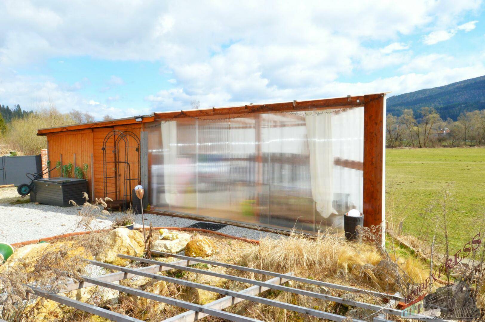 Hölzerner Anbau mit transparenten Wänden und einem Gerüst im Vordergrund, umgeben von ländlicher Landschaft.