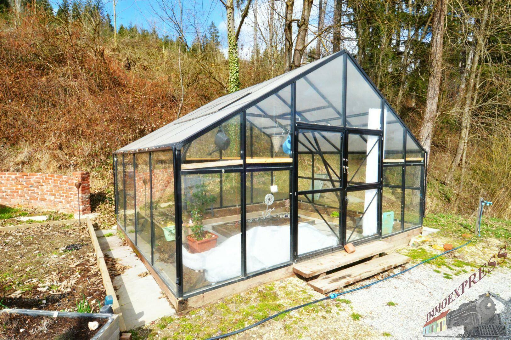 Modernes Gewächshaus mit Glaswänden und schwarzem Rahmen, umgeben von Gartenfläche und Bäumen.