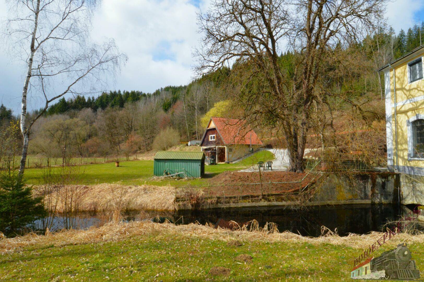 Ländliche Szenerie mit einem gelben Gebäude, einem kleinen Teich und einem weiteren Haus im Hintergrund, umgeben von Natur.