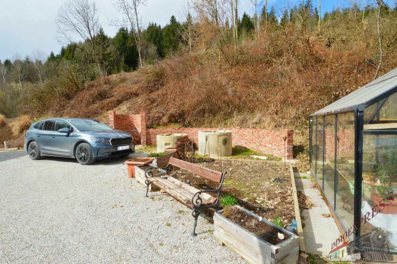 Kiesparkplatz mit einem Auto, einer Gartenbank und einem Gewächshaus vor einer bewaldeten Anhöhe.