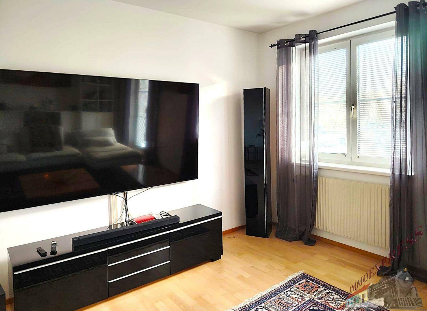 Moderner Wohnbereich mit großem Flachbildfernseher, dunklen Vorhängen und hellem Holzboden.