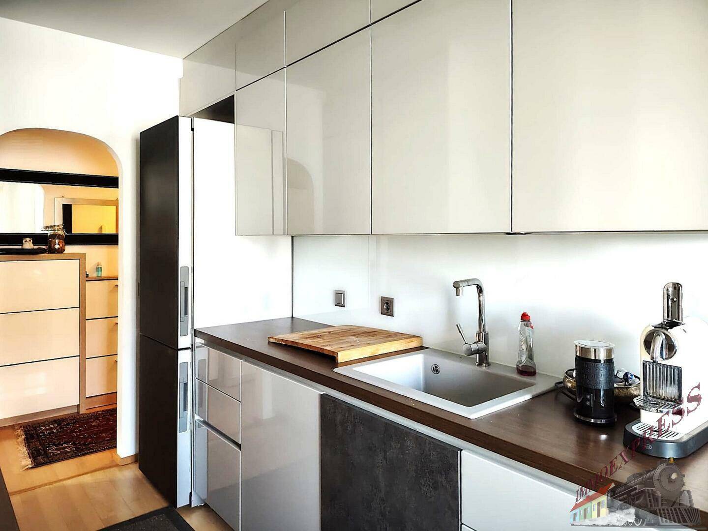 Moderne Küche mit glänzenden weißen Schränken, Spüle und Kaffeemaschine, offen zum Flur.