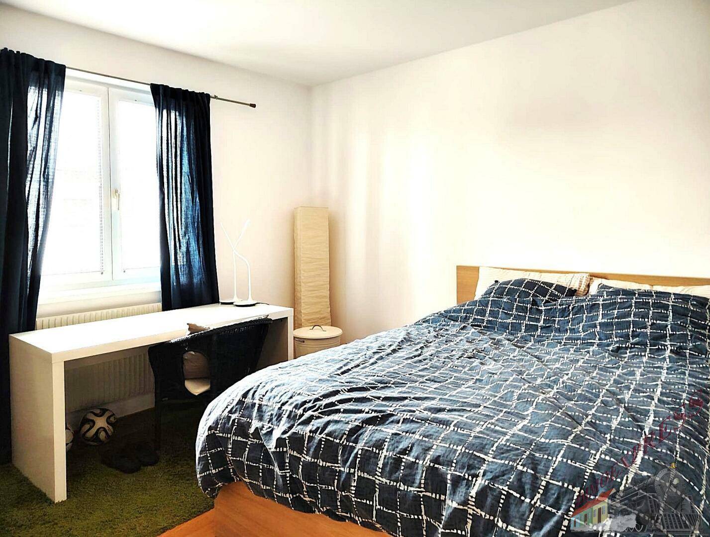 Helles Schlafzimmer mit Bett, Schreibtisch und Fenster mit blauen Vorhängen.