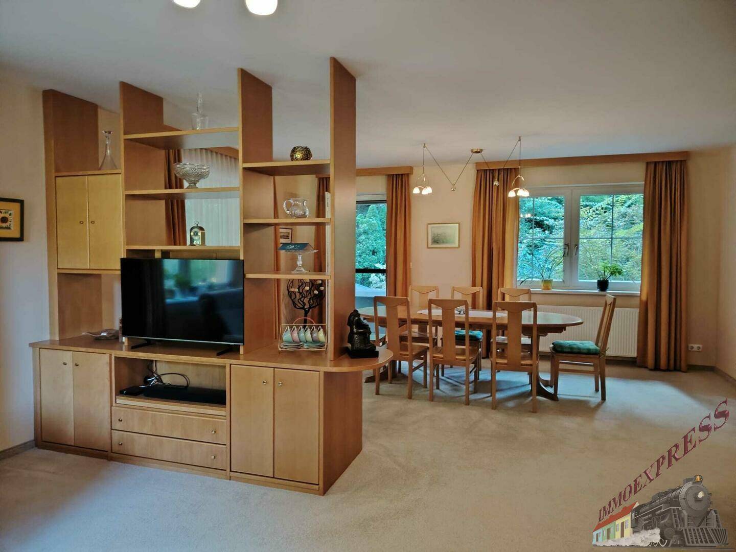Geräumiger Wohn- und Essbereich mit Teppichboden, Holzmöbeln und einem Raumteiler mit Fernseher.