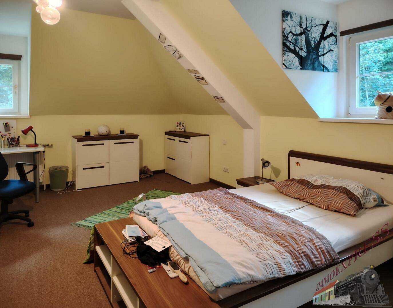 Helles Schlafzimmer im Dachgeschoss mit schrägen Wänden, Teppichboden und einem Fenster mit Grünblick.