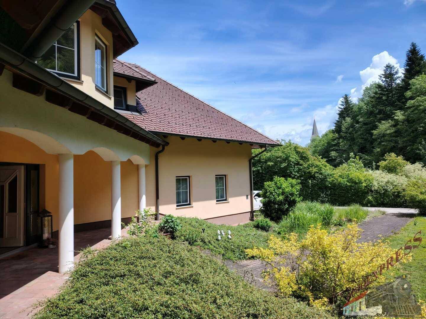 Ansicht des Hauses mit Säulenportal und Blick auf den weitläufigen Garten und die bewaldete Umgebung.