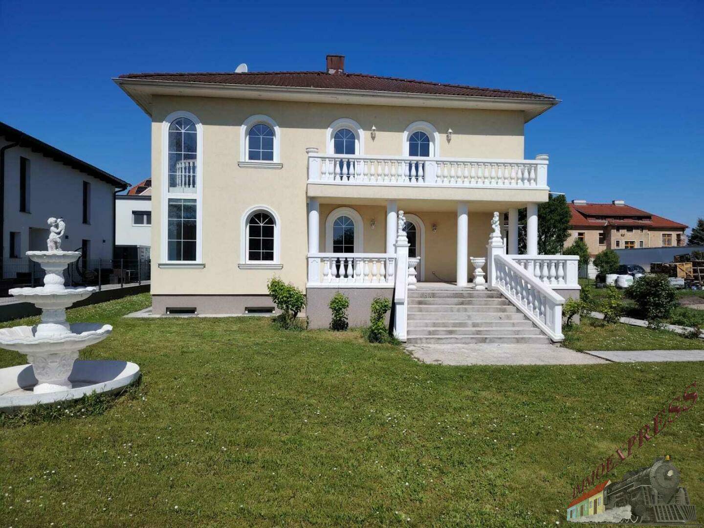 Herrschaftliche Villa mit großem Rasen und dekorativem Brunnen im Vorgarten bei strahlendem Sonnenschein.
