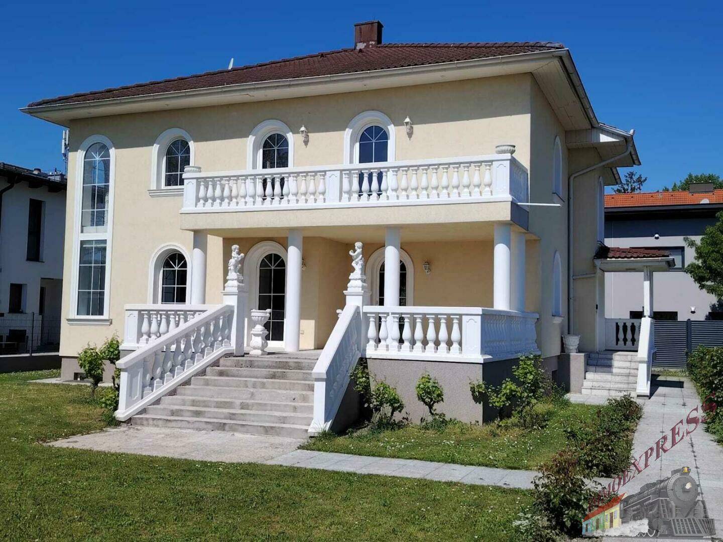 Klassische Villa mit hellem Anstrich, Säulenbalkon und gepflegtem Vorgarten an einem sonnigen Tag.