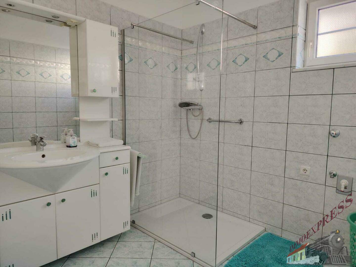 Badezimmer mit Dusche, Waschbecken und Fenster, ausgestattet mit hellen Fliesen und Haltegriffen.