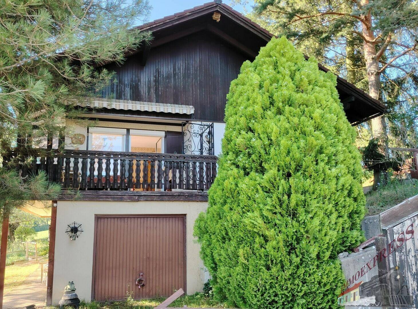 Vorderansicht des Hauses mit integrierter Garage, Holzbalkon und üppiger Bepflanzung.