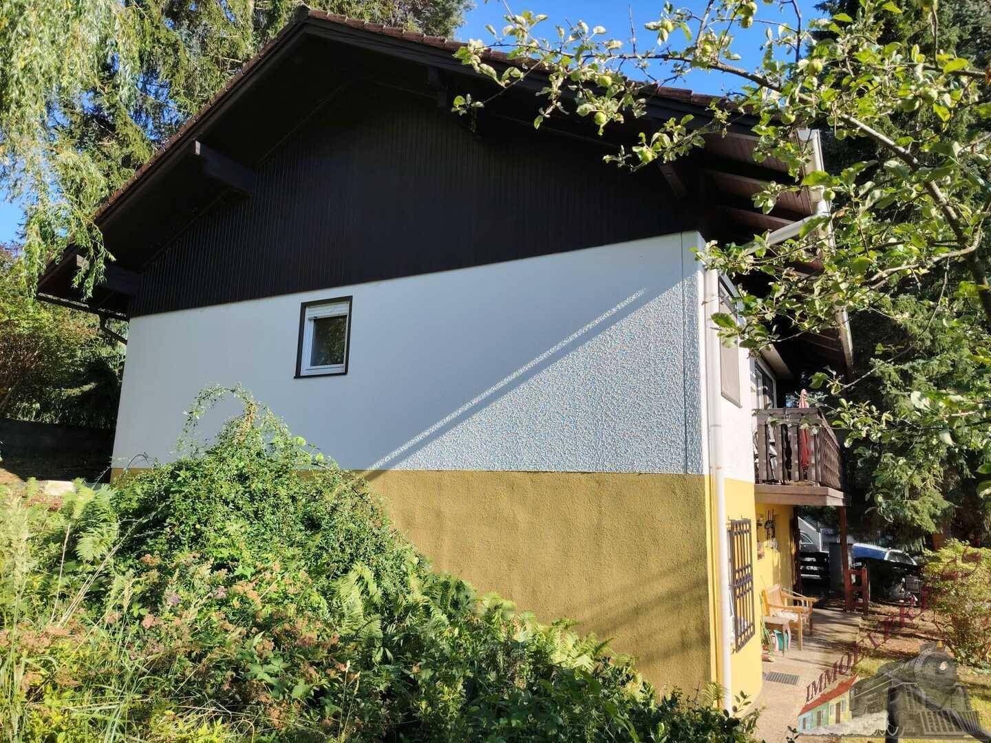 Seitenfassade des Hauses mit gelb-weißem Putz und dunklem Holzdach, umgeben von Grün.