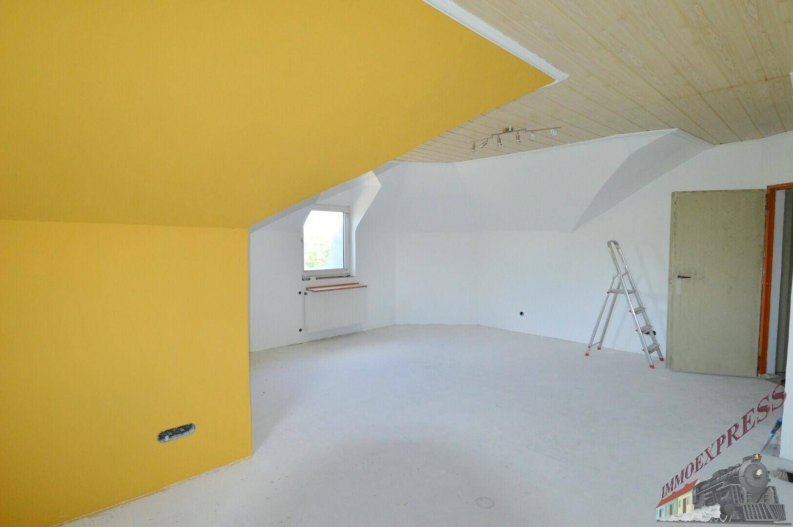 Unfertiger Dachbodenraum mit gelben Wänden, schrägen Decken und einem Fenster, bereit für den Ausbau.