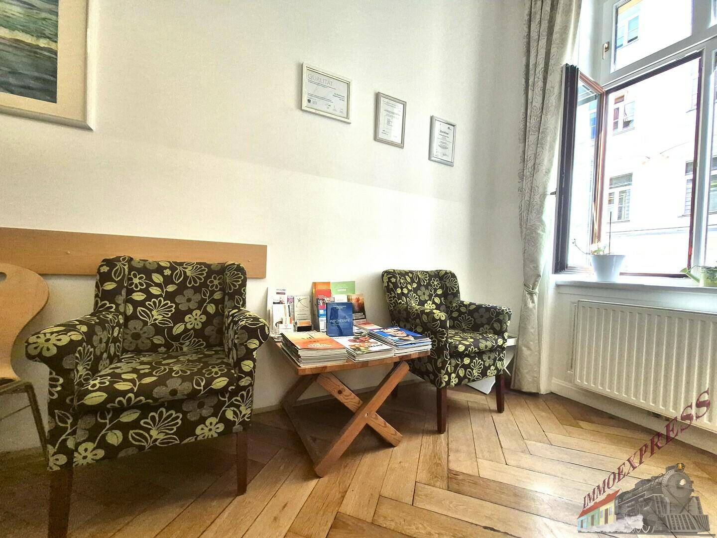 Detailansicht des Wartebereichs mit floralen Sesseln, einem Beistelltisch und einem geöffneten Fenster.