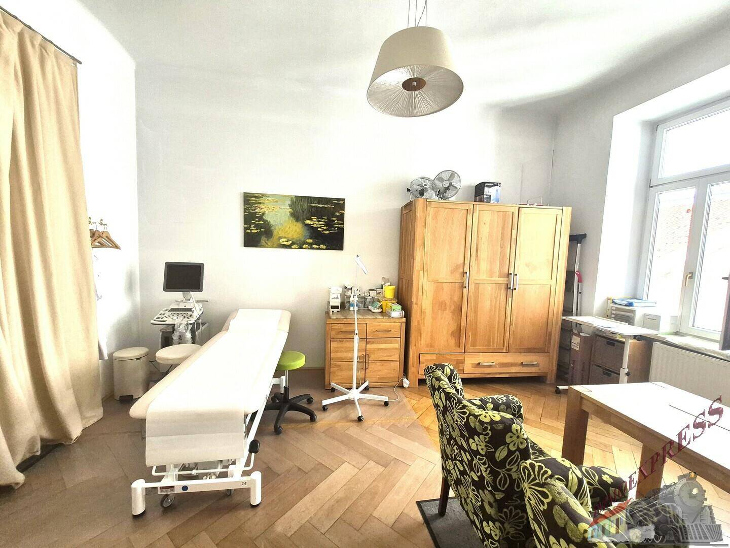 Geräumiger Praxisraum mit Untersuchungsbank, Holzschrank und floral gemustertem Sessel, hell durch großes Fenster.