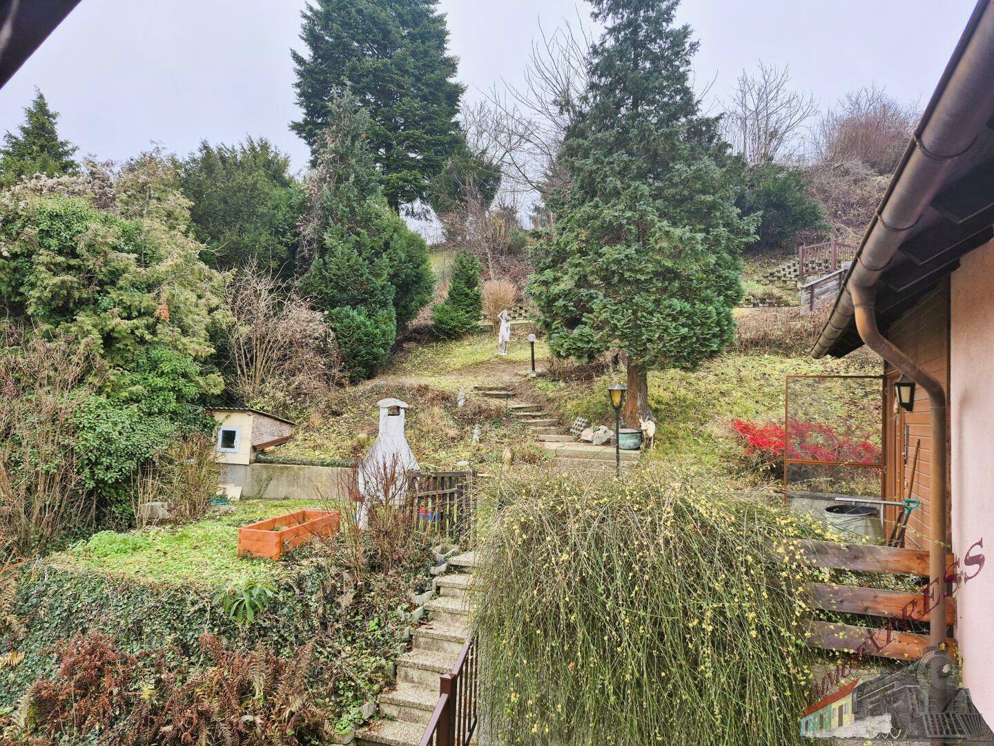 Hanglage-Garten mit Treppen, einem Grillplatz und vielfältiger Vegetation unter bewölktem Himmel.