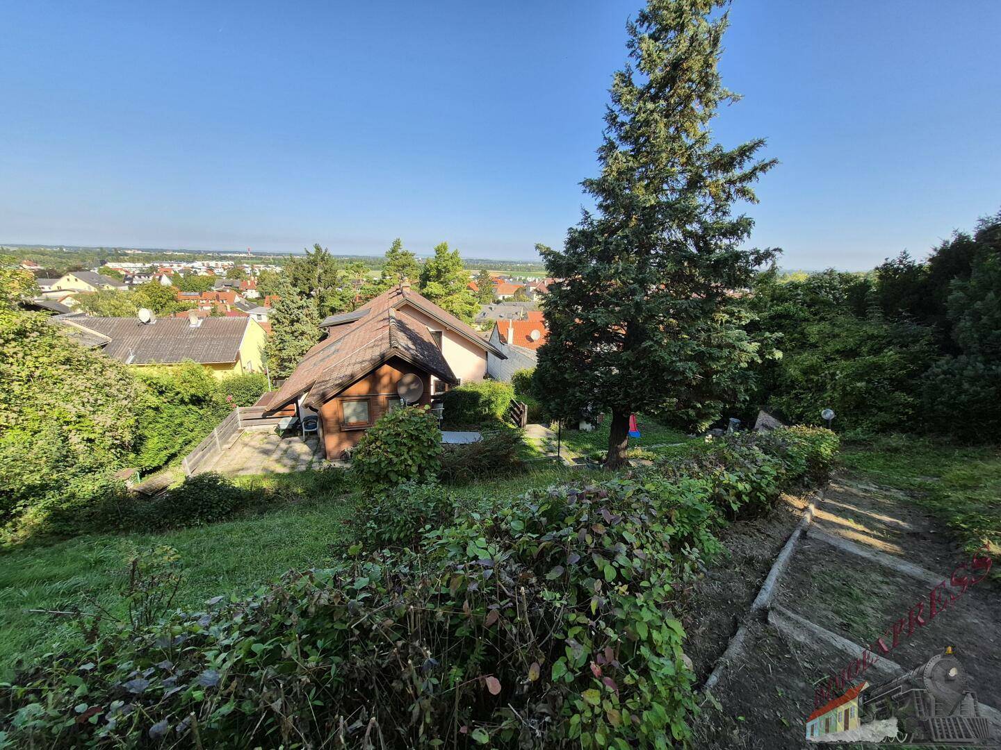 Erhöhte Ansicht eines Hauses mit Ziegeldach, umgeben von Bäumen und Blick auf eine Ortschaft.