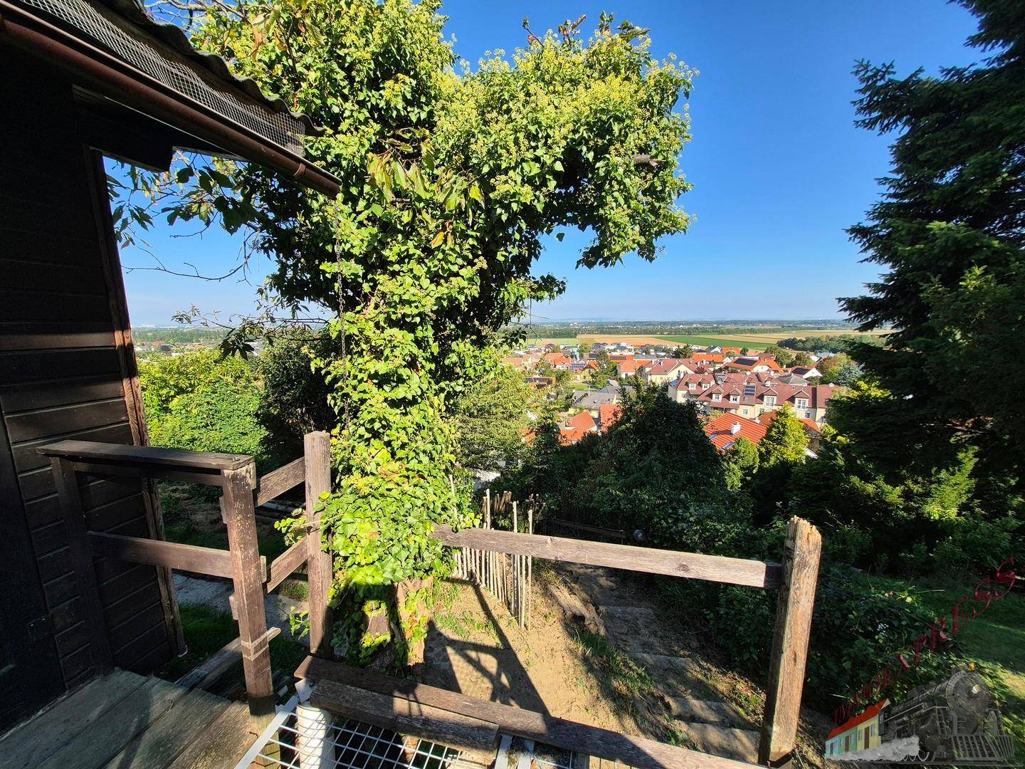 Aussicht von einer Holzkonstruktion auf eine sonnige Ortschaft, umgeben von üppigem Grün.