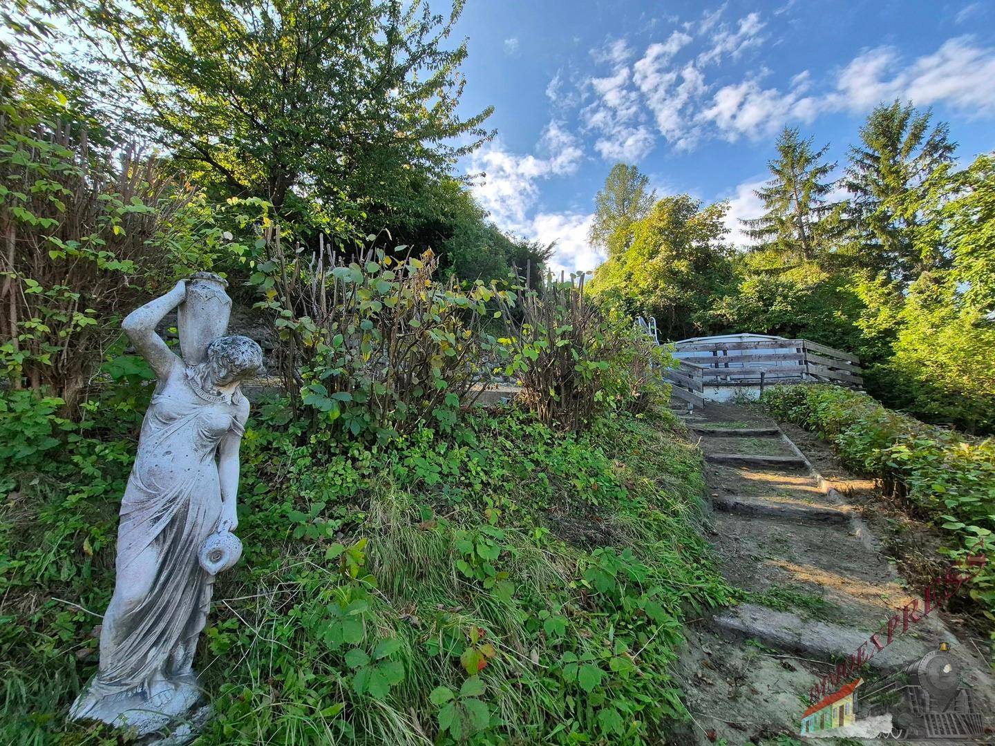 Grüner Hanggarten mit klassischer Statue, Treppen und einem Poolbereich in der Ferne.