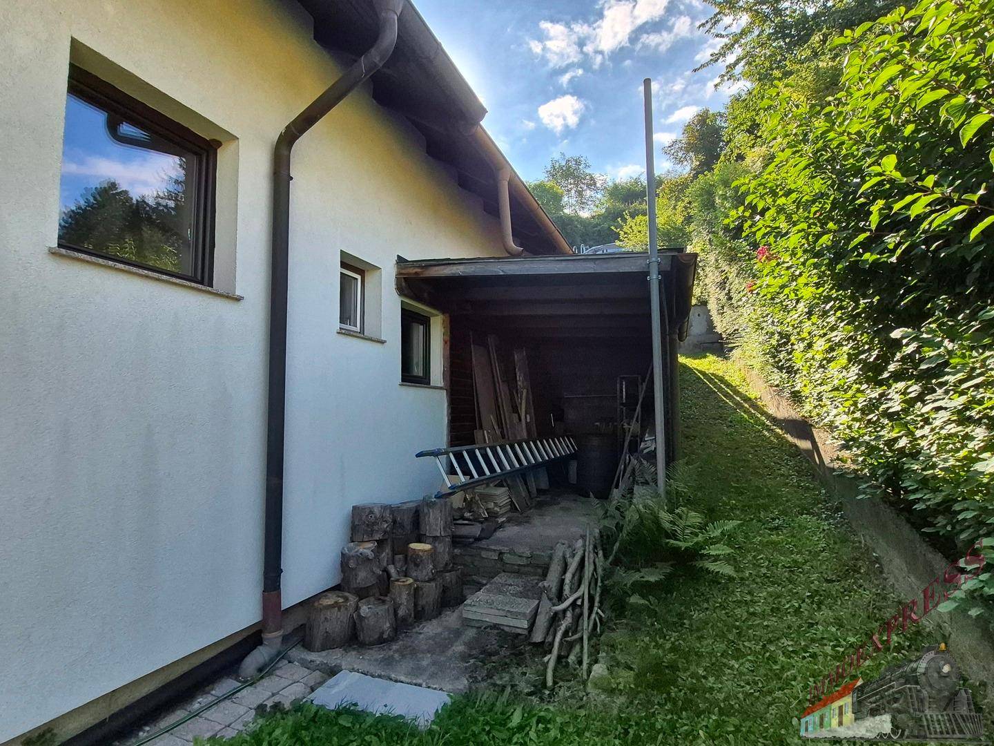 Seitenansicht des Hauses mit überdachtem Lagerbereich und bewachsenem Hang.