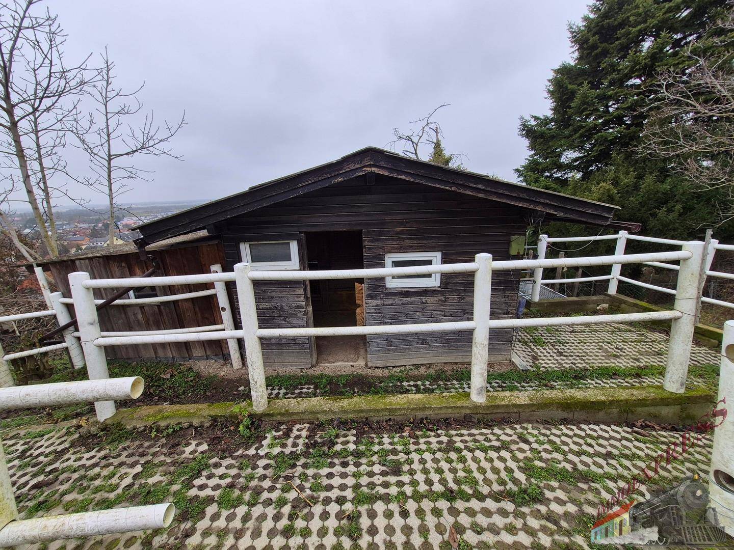 Kleines Holzhaus oder Gartenhütte mit weißem Zaun auf einem gepflasterten Hanggrundstück.