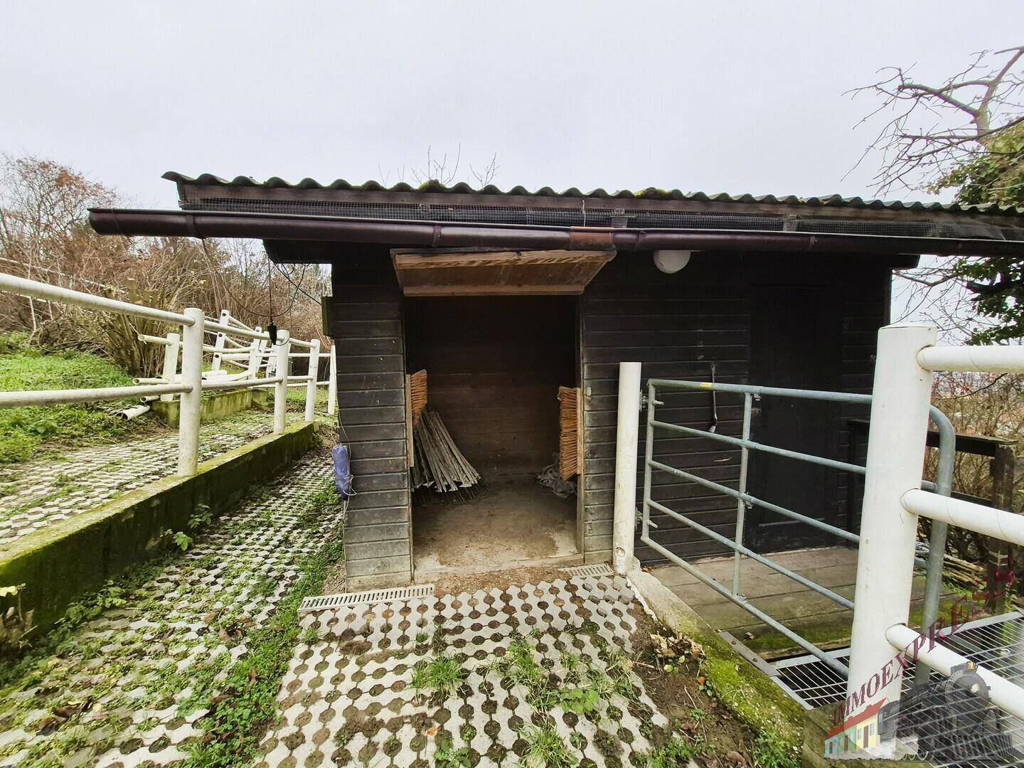Seitenansicht der Holzhütte mit offenem Eingang und Metalltor auf dem gepflasterten Gelände.