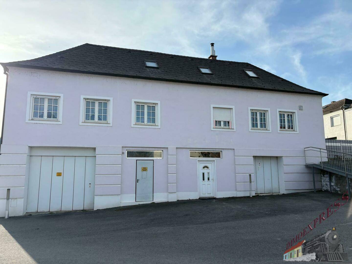 Vorderansicht des Gebäudes mit Garagentoren, Eingangstüren, Fenstern und einer Außentreppe auf der rechten Seite.