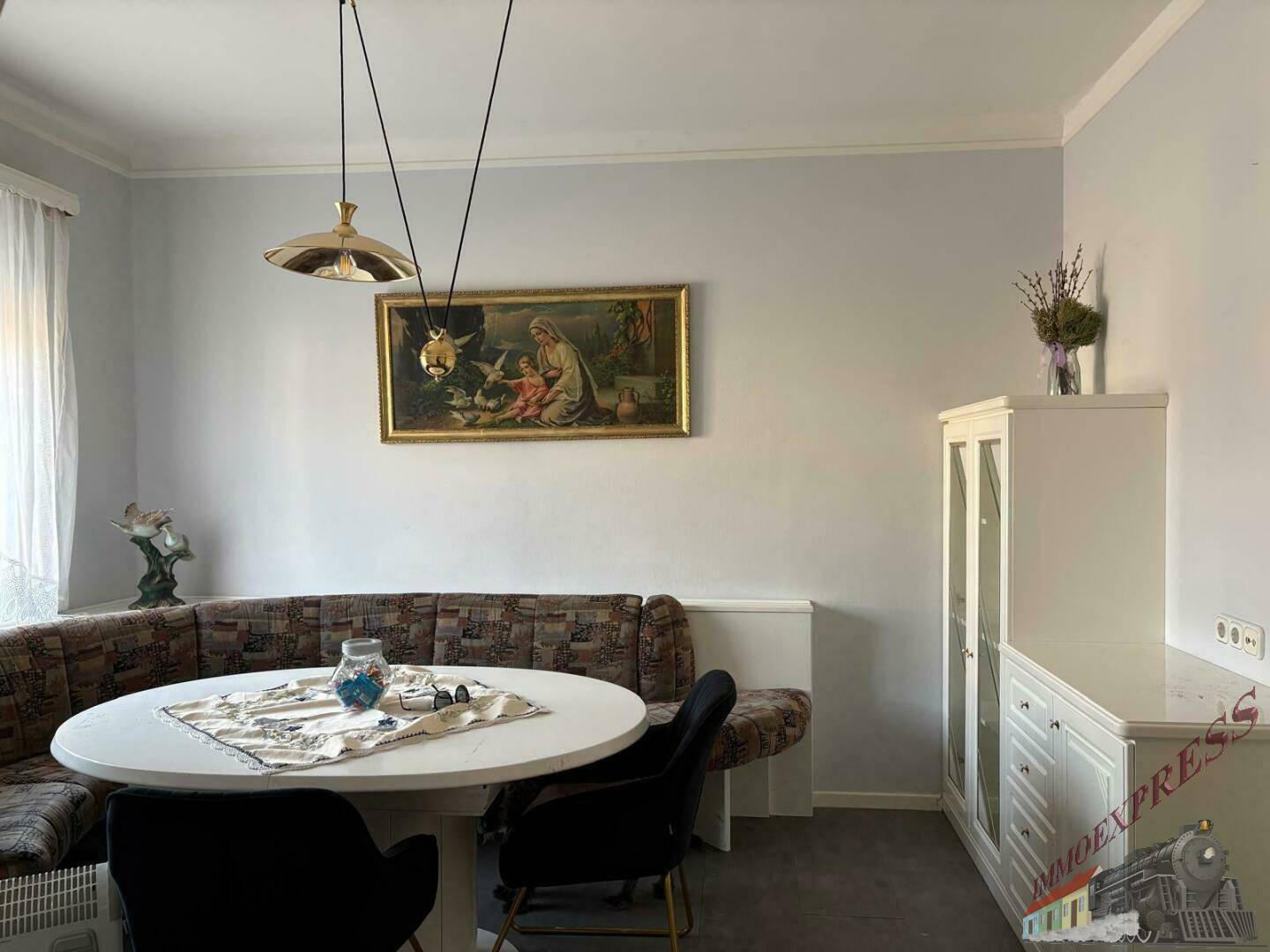 Essbereich mit rundem weißen Tisch, Eckbank, Stühlen und einem weißen Schrank, unter hellgrauen Wänden.