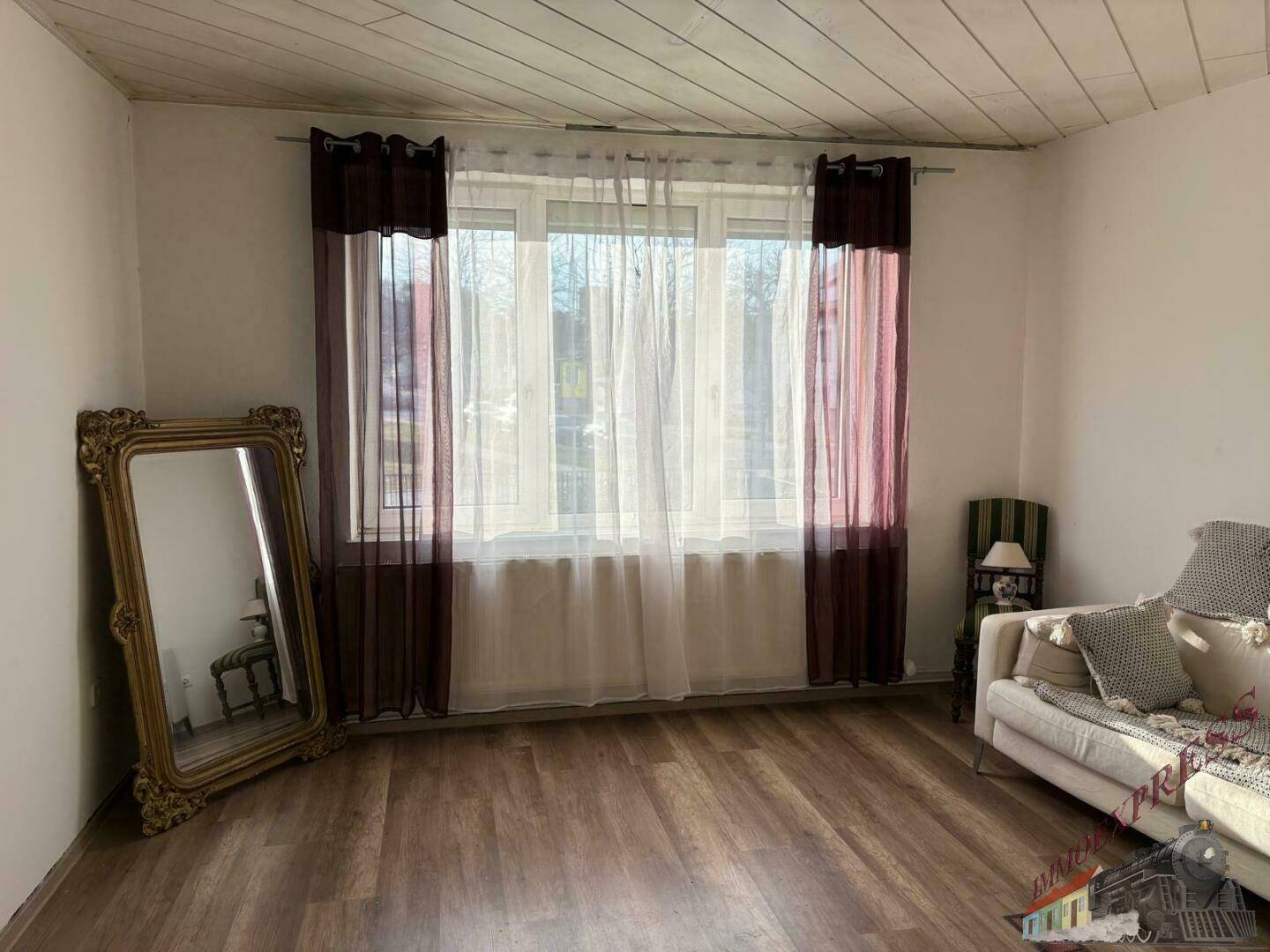 Heller Wohnraum mit großem Fenster, Vorhängen, einem dekorativen Spiegel und Laminatboden unter einer Holzdecke.