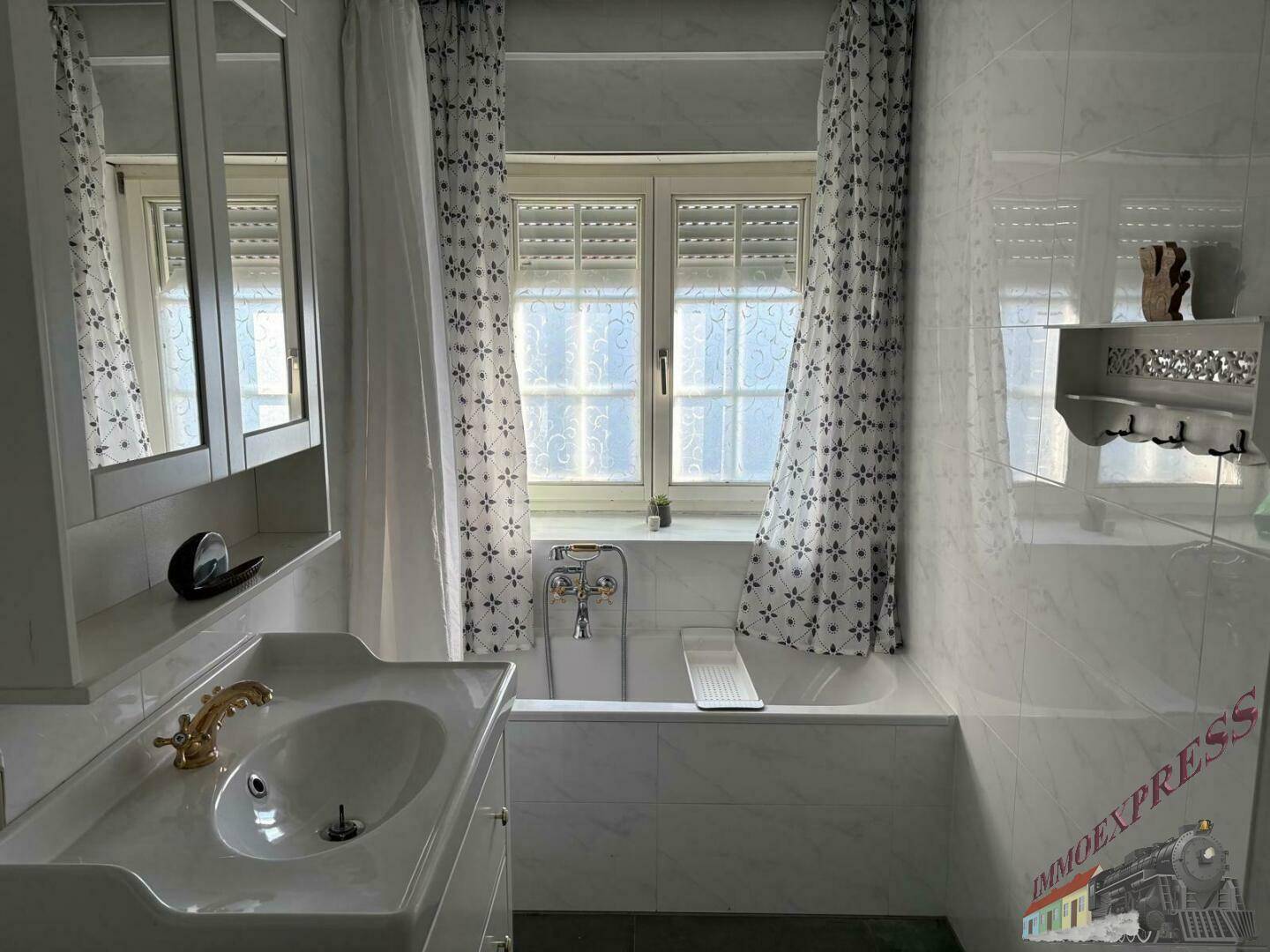 Badezimmer mit Badewanne, Waschtisch, Spiegelschrank und gefliesten Wänden und Böden, mit Fenster und Rollläden.