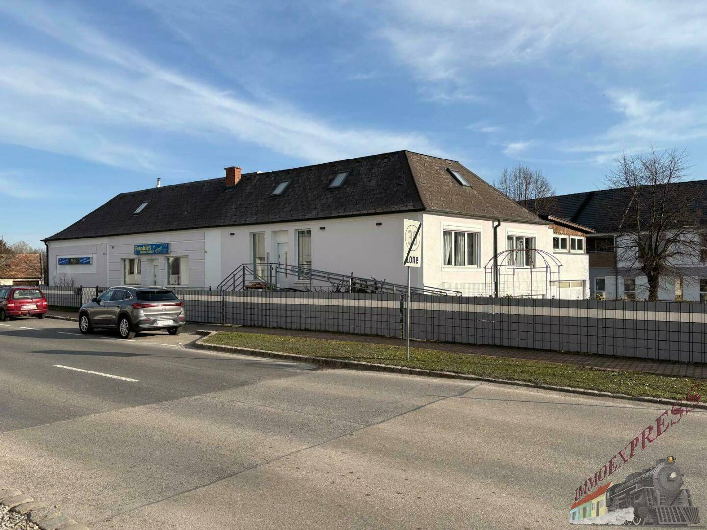 Langes, weißes Gebäude mit dunklem Dach, Fenstern und einer Rampe, gelegen an einer Straße mit parkenden Autos.