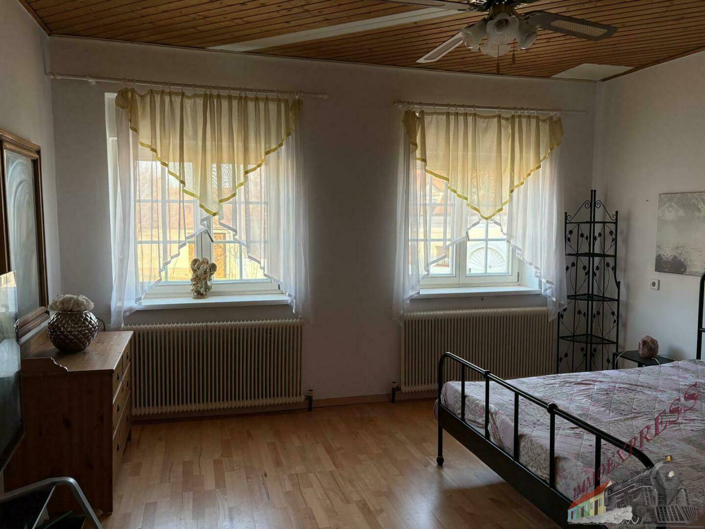 Schlafzimmer mit Bett, Kommode, zwei Fenstern mit Vorhängen und Holzfußboden unter einer Holzpaneeldecke.