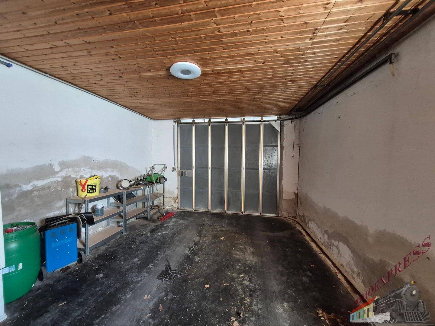 Garage mit Holzbalkendecke, Betonboden und einem großen Rolltor am Ende des Raumes.